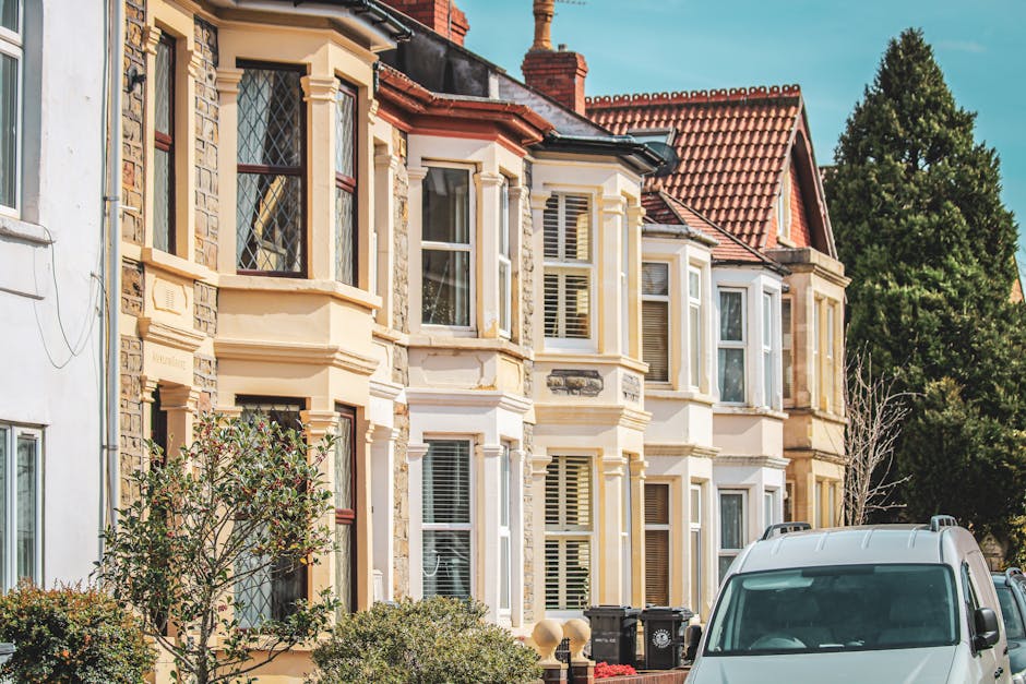 Photograph of a row of multi-storey residential buildings with bay windows, decorative stonework, and red-tiled roofs, situated on a street with trees and greenery in the background. In the foreground