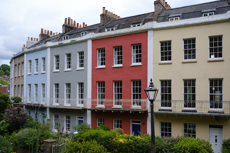 Photograph of a row of multi-storey residential buildings with bay windows, decorative stonework, and red-tiled roofs, situated on a street with trees and greenery in the background. In the foreground
